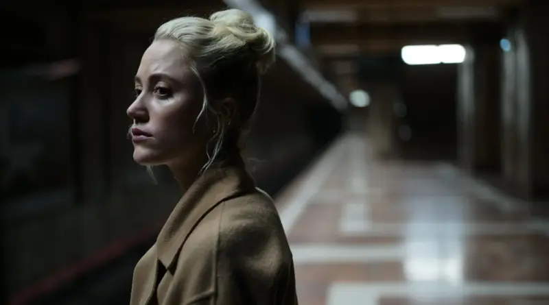 Fotograma De La Película 'El Extraño'. Plano Medio Lateral De Una Mujer Joven Rubia (Maika Monroe) Con El Pelo Recogido En Un Moño Alto Y Un Abrigo De Lana Beige. Está De Pie En Una Estación De Metro O Pasillo Subterráneo Con El Fondo Desenfocado. Mira Hacia Su Derecha Con Una Expresión De Alerta, Tensión Y Preocupación, Como Si Hubiera Escuchado O Visto Algo Sospechoso.