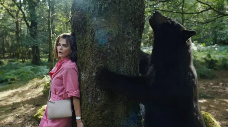 Keri Russell escondida tras un árbol mientras un oso negro busca algo al otro lado en Oso Vicioso.