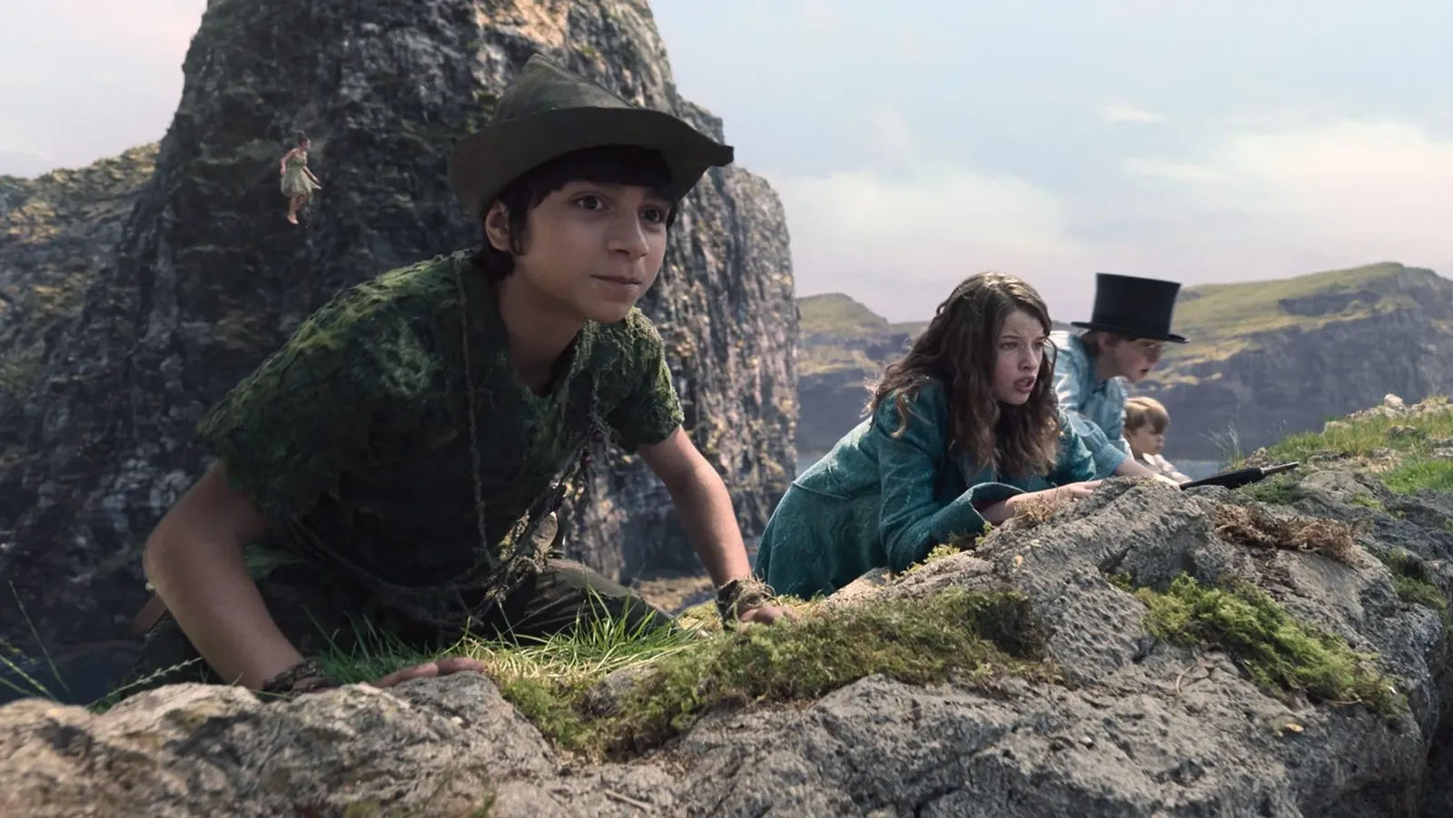 Fotograma de la película 'Peter Pan & Wendy'. En primer plano, Peter Pan aparece agachado sobre una roca cubierta de musgo, vistiendo su túnica verde de texturas naturales y un sombrero puntiagudo. A su lado, Wendy, con un camisón azul, y los niños perdidos observan con cautela desde lo alto de un acantilado. Al fondo, se erigen grandes formaciones rocosas sobre un mar en calma bajo un cielo nublado, con la pequeña figura de Campanilla volando cerca de las rocas.