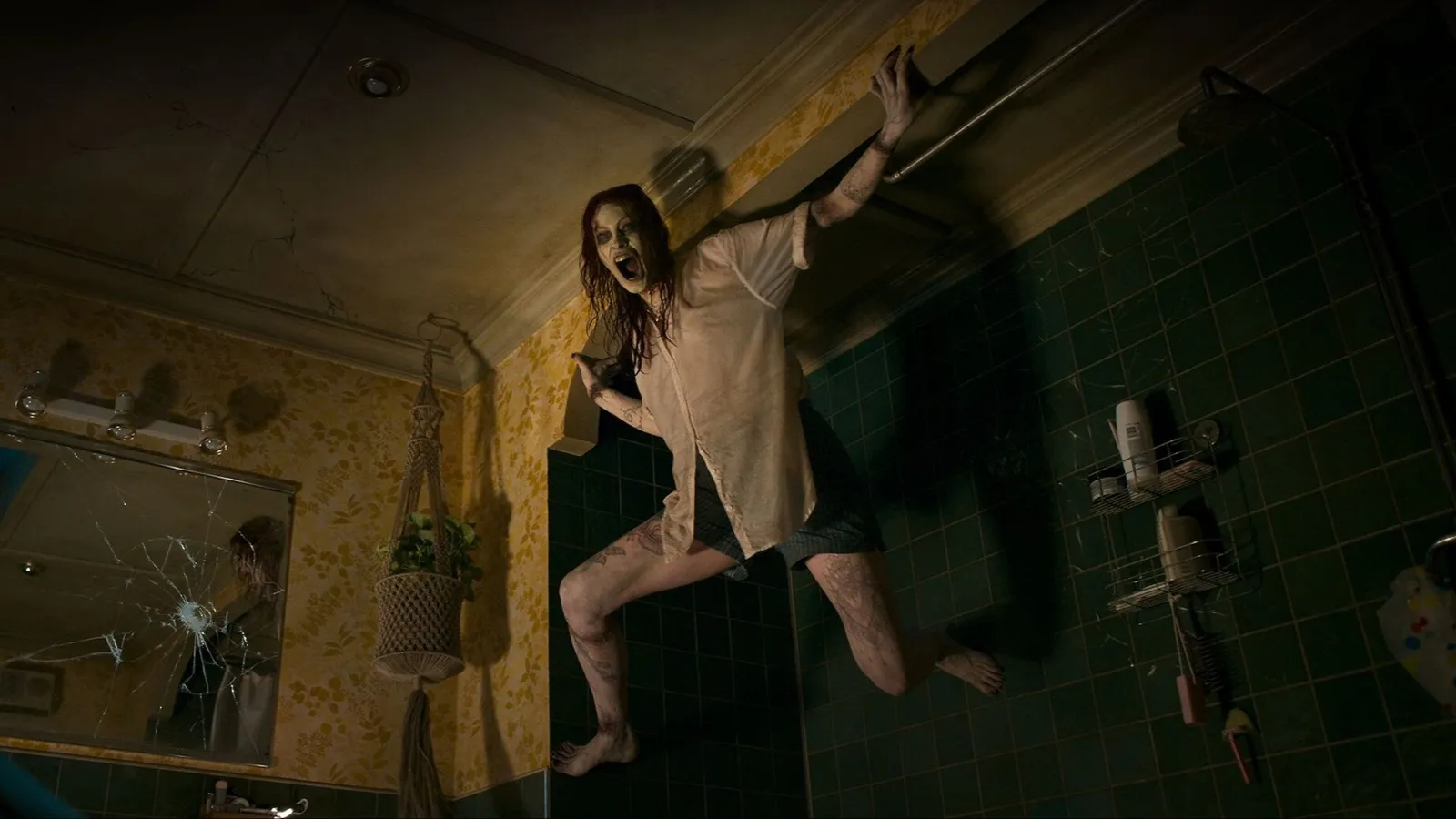 Fotograma de la película 'Posesión infernal: El despertar'. Una mujer poseída, con el rostro pálido y desencajado, el cabello rojo desordenado y una expresión de grito aterrador, aparece trepando de forma antinatural por las paredes de un cuarto de baño. Está suspendida en una esquina, apoyada contra azulejos verdes oscuros. Viste una camisa blanca desabrochada y pantalones cortos. A la izquierda, un espejo roto refleja la escena y el papel pintado con motivos florales amarillos de la habitación.