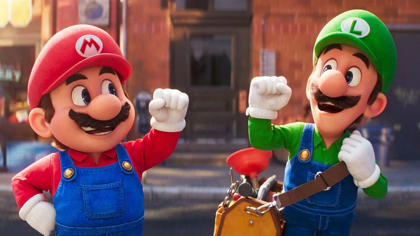 Fotograma de 'Super Mario Bros. La Película' que muestra a Mario (izquierda) y Luigi (derecha) en una calle de Brooklyn. Ambos visten sus icónicos petos vaqueros azules y camisetas de color rojo y verde respectivamente, con sus gorras a juego. Aparecen con una expresión de triunfo y determinación, alzando un puño enguantado en el aire. Luigi lleva al hombro una bolsa de herramientas de fontanero con un desatascador visible. El fondo muestra una calle urbana con edificios de ladrillo desenfocados.