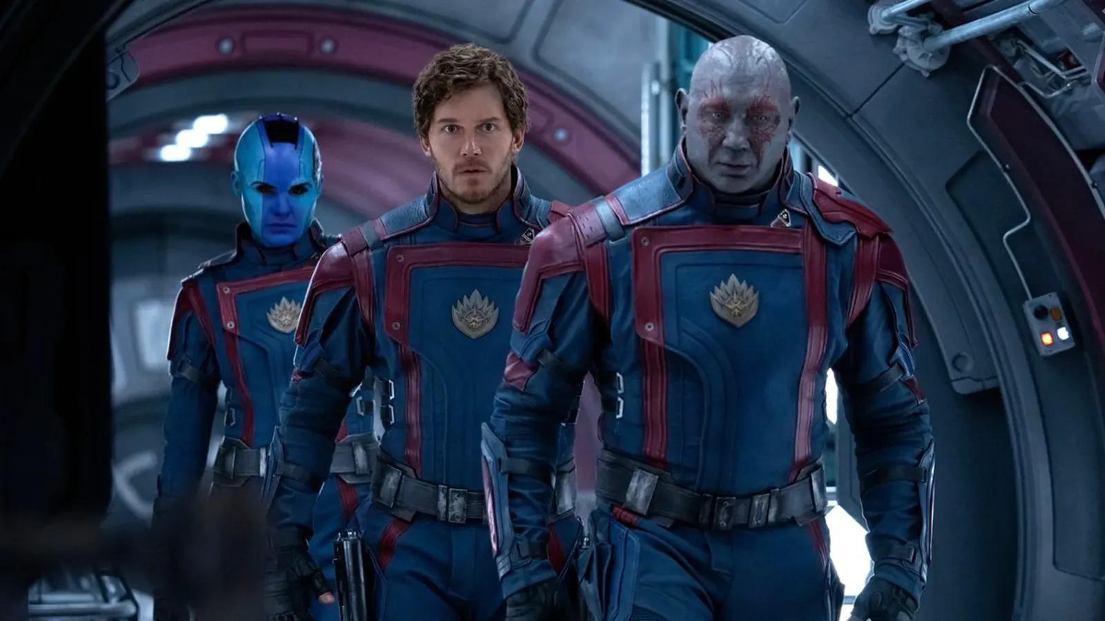 Fotograma de 'Guardianes de la Galaxia Vol. 3' que muestra a Nebula, Peter Quill (Star-Lord) y Drax caminando por el pasillo metálico de una nave espacial. Los tres visten los nuevos uniformes del equipo: trajes tácticos de color azul oscuro con detalles en rojo y el emblema dorado de los Guardianes en el pecho. Nebula tiene la piel azul y mejoras cibernéticas; Peter Quill, en el centro, tiene una expresión de preocupación; y Drax, a la derecha, muestra sus características marcas rojas en la piel gris. La iluminación es fría y proviene de paneles integrados en las paredes curvas.