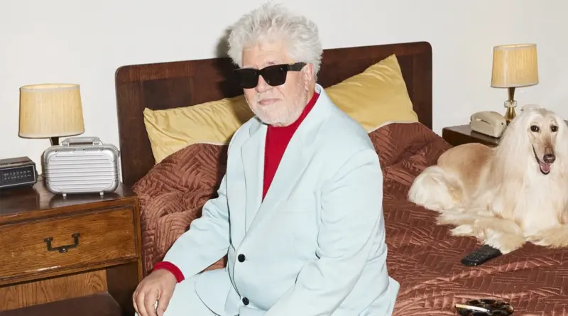 El director de cine Pedro Almodóvar sentado en una cama con traje azul claro y gafas de sol.