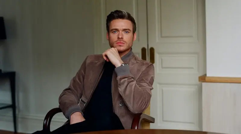 Retrato de Richard Madden sentado, vistiendo una chaqueta marrón y mirando fijamente a cámara.