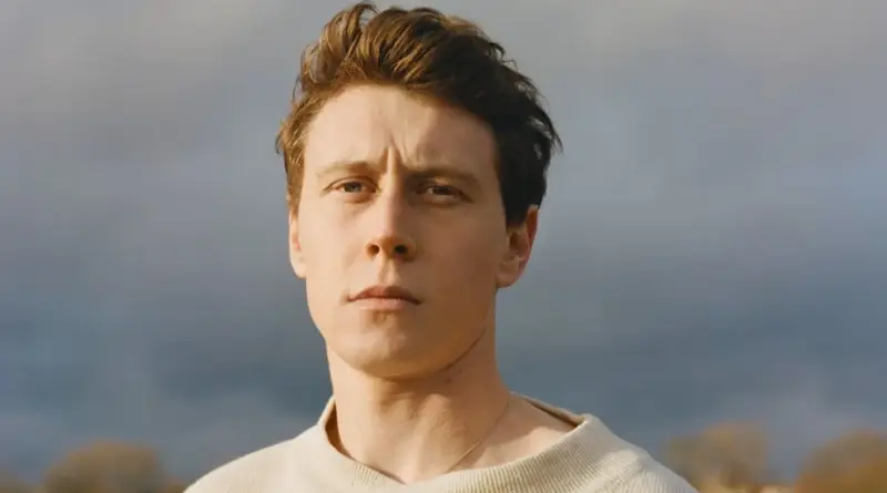 Primer plano del actor George MacKay con suéter beige bajo un cielo nublado.