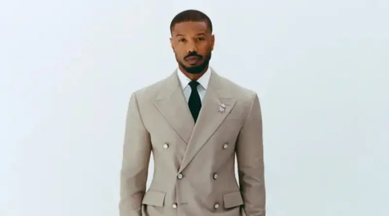 Michael B. Jordan posando con un traje cruzado color beige y corbata negra sobre fondo blanco.