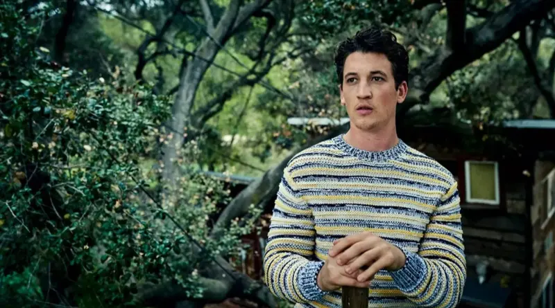 El actor Miles Teller con un suéter de rayas amarillas y azules en un entorno boscoso.