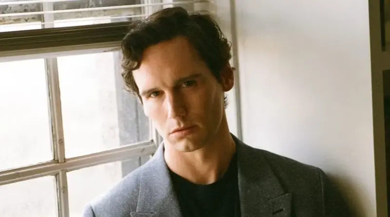 Retrato del actor Cory Michael Smith apoyado en una ventana.