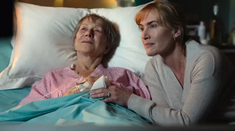 Kate Winslet en 'Adiós, June' sentada junto a la cama de un hospital, sosteniendo la mano de Helen Mirren, quien permanece recostada con una expresión de serenidad.