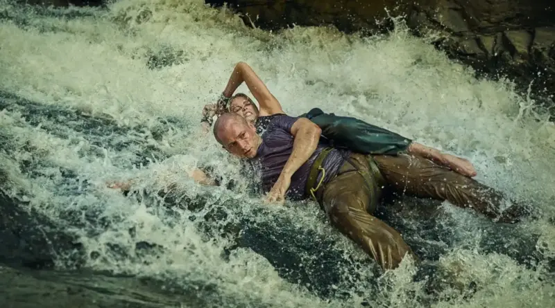 Escena de acción de la película Apex que muestra a Charlize Theron y Taron Egerton luchando por su vida en mitad de unos rápidos de agua embravecida. Ella aparece agarrada a la espalda de él mientras ambos son arrastrados por la fuerte corriente entre rocas.