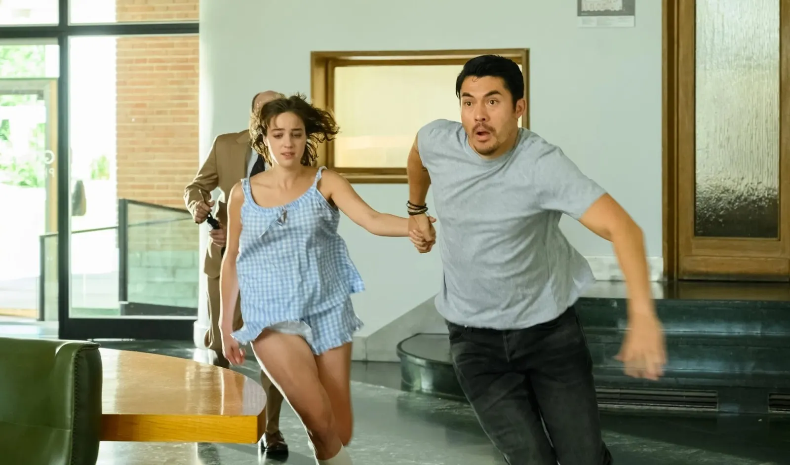 Fotografía de la película Daniela Forever que muestra a los actores Henry Golding y Beatrice Grannò corriendo de la mano a través del vestíbulo de un edificio moderno. Él viste una camiseta gris y pantalones negros con expresión de asombro, mientras ella luce un pijama de cuadros azules y blancos. Al fondo, un hombre con traje marrón intenta alcanzarlos.