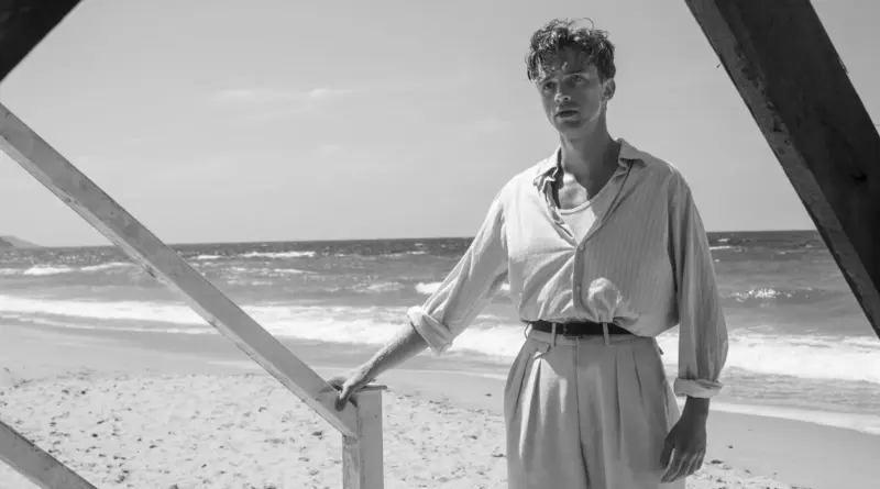 Fotografía en blanco y negro del actor Benjamin Voisin en la película 'El Extranjero' como Meursault, de pie en una playa soleada junto a una barandilla de madera, vistiendo una camisa de rayas claras y pantalones de talle alto.