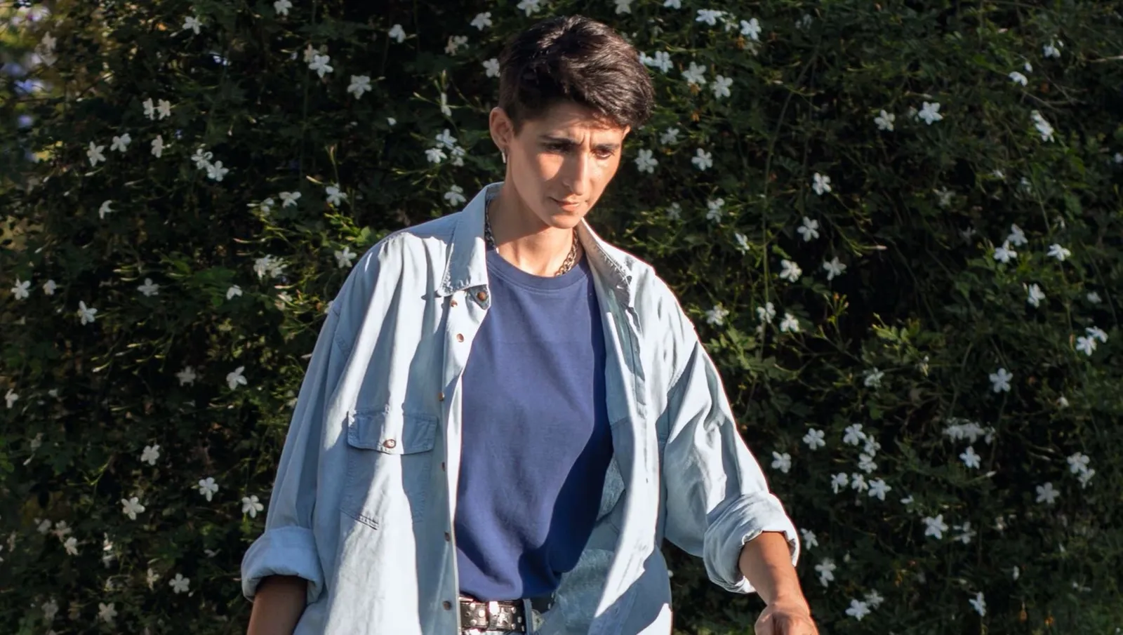 Fotografía del documental Flores para Antonio que muestra a Alba Flores caminando en un entorno exterior. Viste una camiseta azul oscuro bajo una camisa de color azul claro desabrochada, con el cabello corto y una expresión de serena introspección. Al fondo se aprecia un seto denso cubierto de pequeñas flores blancas.