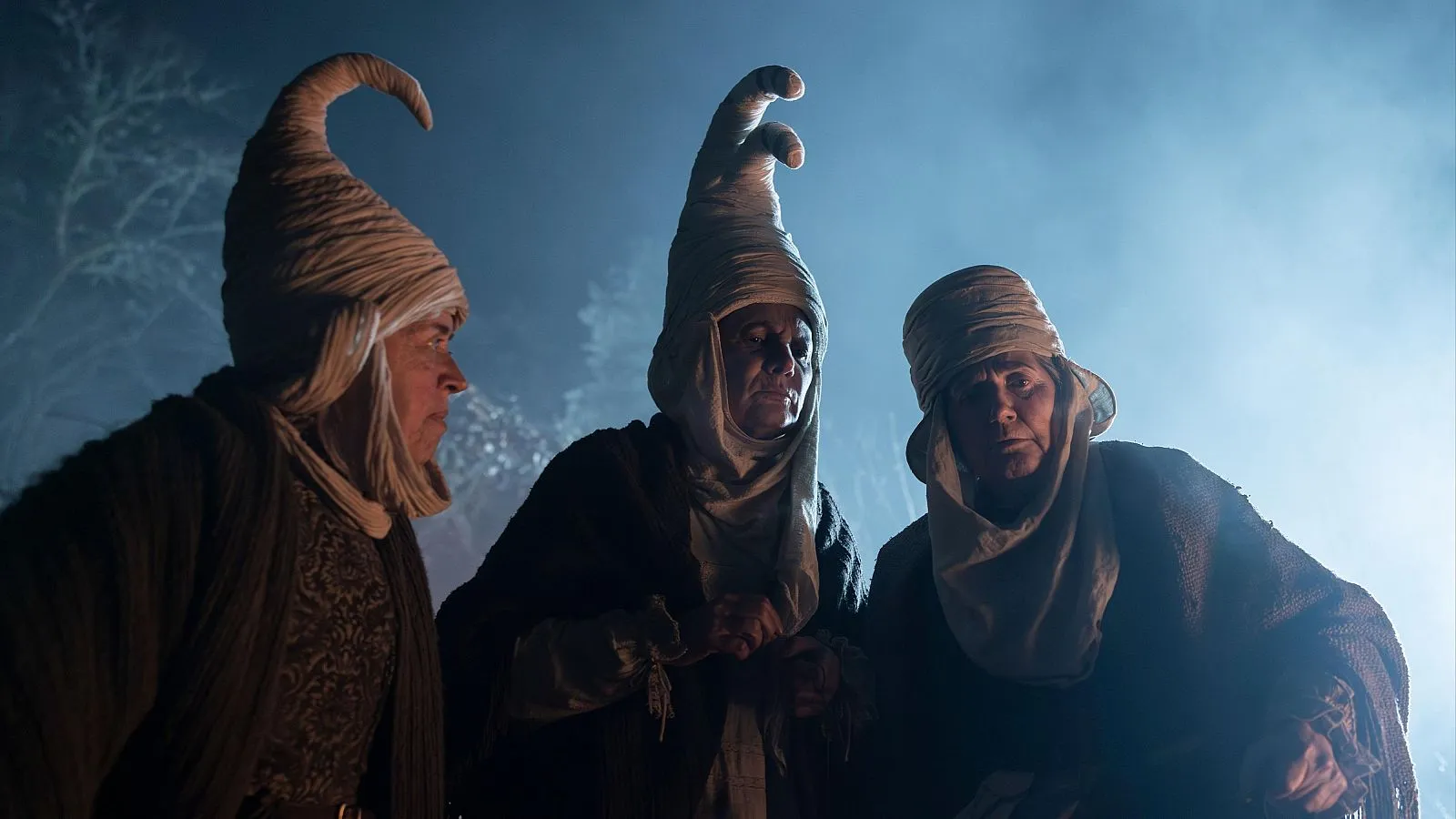otografía de la película Gaua (2025) de Paul Urkijo Alijo que muestra a las actrices Elena Irureta, Ane Gabarain e Iñake Irastorza caracterizadas como tres mujeres del siglo XVII. Visten túnicas oscuras y tocados blancos en forma de cono alargado y curvado que cubren sus cabezas. Están situadas en un exterior nocturno con una iluminación azulada y brumosa que resalta sus rostros de expresión grave y misteriosa.