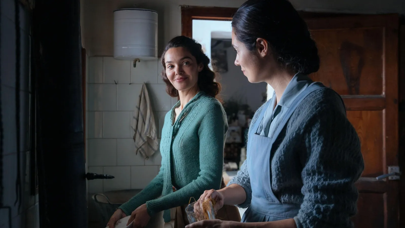 Fotografía de la película La buena letra que muestra a dos mujeres en una cocina de época con paredes de azulejos blancos. Una mujer en primer plano, de perfil, viste un delantal azul y limpia un vaso, mientras que la mujer al fondo, con una rebeca verde, la observa con una sonrisa sutil y afectuosa. La iluminación es naturalista, filtrándose desde un lateral.