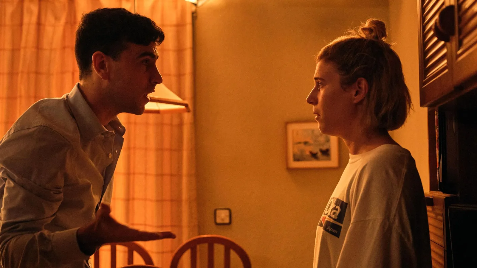 Fotografía de la película La furia que muestra a un hombre, Alex Monner, y una mujer, Ángela Cervantes, enfrentados en una habitación con luz cálida y anaranjada. El hombre, a la izquierda, viste una camisa clara y gesticula con la mano abierta mientras habla con intensidad. La mujer, a la derecha, viste una camiseta blanca con letras impresas, lleva el pelo recogido en un moño y mantiene una expresión tensa y de escucha seria. Al fondo se aprecia una cortina de cuadros y un pequeño cuadro en la pared.