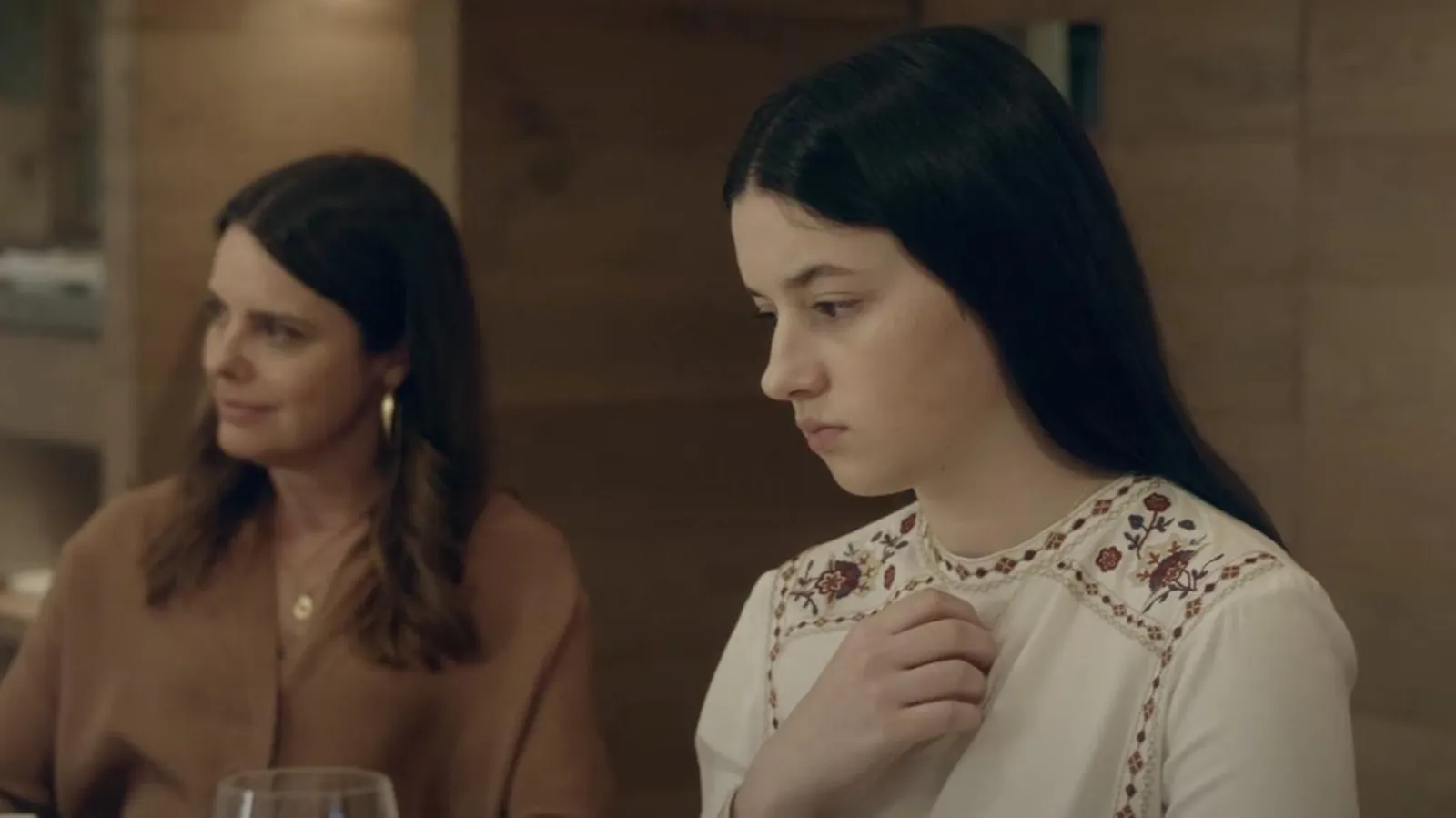 Fotografía de la película Los domingos que muestra a una mujer joven, Blanca Soroa, en primer plano, de perfil, con una expresión pensativa y melancólica. Viste una blusa blanca con bordados florales en los hombros y tiene el cabello largo y oscuro. Al fondo, desenfocada, se observa a una mujer mayor con una blusa marrón y pendientes dorados, mirándola con curiosidad.