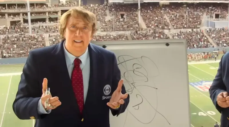 Nicolas Cage caracterizado como John Madden en la película Madden de Amazon MGM Studios, luciendo una prótesis facial, cabello rubio peinado hacia un lado y un traje azul marino con el logo de CBS Sports. El actor aparece en una cabina de retransmisión frente a una pizarra blanca con garabatos tácticos y un estadio de la NFL lleno de público al fondo.