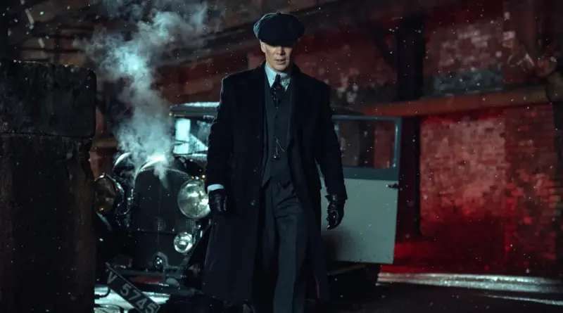 El actor Cillian Murphy como Tommy Shelby en 'Peaky Blinders: El Hombre Inmortal', vistiendo su abrigo largo oscuro y gorra característica, camina hacia adelante en una calle nocturna nevada frente a un coche antiguo accidentado.