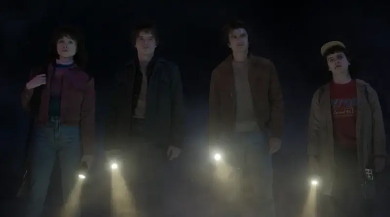 Fotograma de 'Stranger Things 5' que muestra a Nancy, Jonathan, Steve y Dustin en un entorno oscuro y neblinoso. Los cuatro personajes están de pie, mirando hacia arriba con expresiones de asombro y preocupación mientras sostienen linternas encendidas que apuntan hacia el suelo, creando haces de luz entre la bruma. Visten sus ropas características de los años 80: Nancy con una chaqueta granate, Jonathan con una oscura, Steve con una marrón y Dustin con su gorra y una camiseta roja.