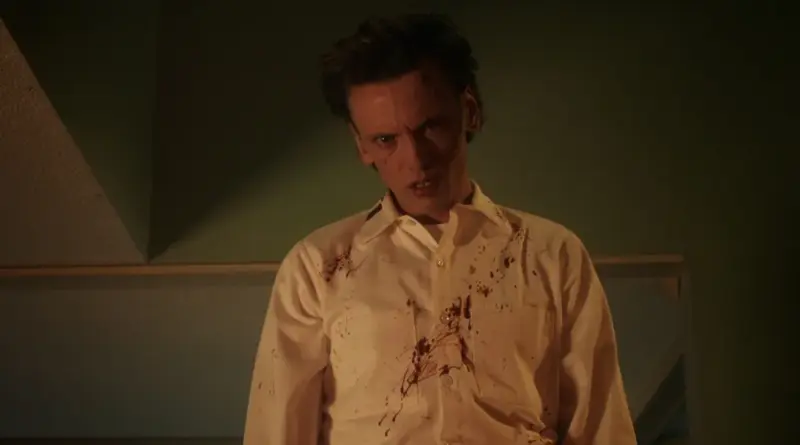 El actor Jamie Campbell Bower como Henry Creel de 'Stranger Things', vistiendo una camisa blanca manchada de sangre y mirando fijamente con expresión perturbada en una habitación con paredes verdes.