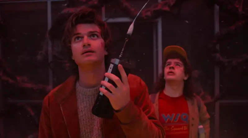 Imagen de la serie 'Stranger Things' que muestra a Steve Harrington en primer plano sosteniendo un walkie-talkie con una expresión de preocupación, mientras Dustin Henderson mira hacia arriba asombrado al fondo. La escena está iluminada con tonos rojos intensos y rodeada de partículas suspendidas y raíces oscuras del Mundo del Revés.