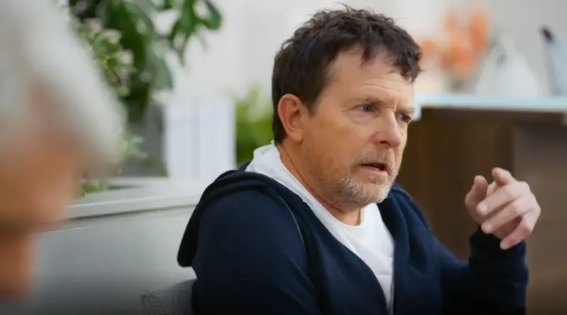 Primer plano de Michael J. Fox en una escena de la tercera temporada de "Terapia sin filtro" (Shrinking) de Apple TV. El actor, con el cabello castaño alborotado y barba de pocos días, viste una sudadera azul marino sobre una camiseta blanca. Mantiene una expresión de escucha atenta y gesticula con su mano derecha mientras mantiene una conversación con Harrison Ford en una sala de espera luminosa.