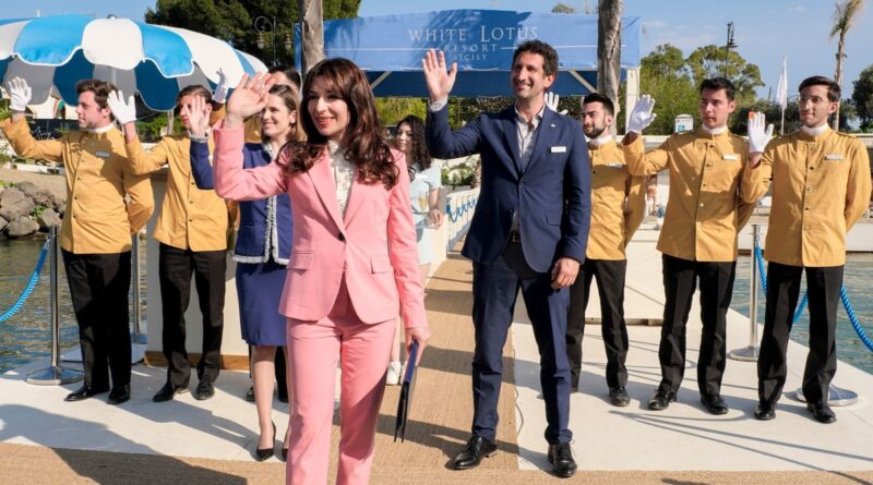 Un grupo de empleados saludando y sonriendo frente a la entrada del 'White Lotus Resort Sicily', de la temporada 2, con palmeras y un cielo despejado de fondo.