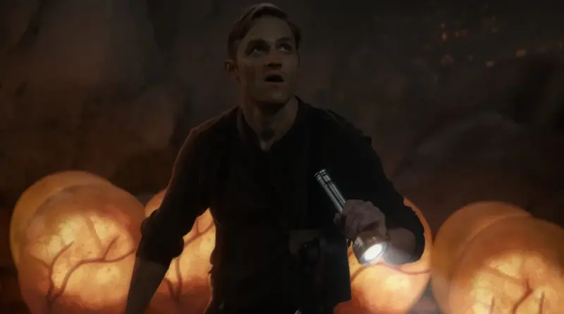 El actor Wyatt Russell, interpretando al joven Lee Shaw en Monarch: El legado de los monstruos, sostiene una linterna con expresión de asombro mientras explora una cueva rodeada de huevos de Titán brillantes.