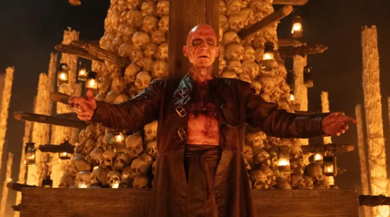 Fotograma de la película '28 Años Después: El Templo de los Huesos'. Ralph Fiennes calvo, demacrado y con la piel marcada o quemada, está de pie con los brazos abiertos en señal de bienvenida o sacrificio. Viste una chaqueta de cuero larga abierta sobre el torso desnudo y sucio. Detrás de él se alza una estructura macabra hecha enteramente de cientos de cráneos humanos apilados, iluminados por linternas colgantes.