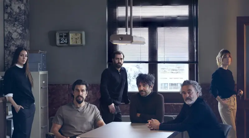 Foto de grupo del reparto de 'En nombre de otro' en un decorado de oficina gris con nieve visible por la ventana. De izquierda a derecha: Blanca Suárez de pie, Mario Casas sentado con camiseta gris, un hombre de pie al fondo, el director Oriol Paulo sentado en el centro, Eduard Fernández sentado a la derecha y Alexandra Jiménez de pie en el extremo derecho. Un reloj en la pared marca "DIC MIE 24".