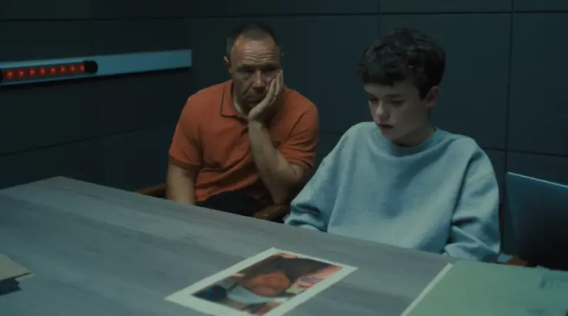 Fotograma de 'Adolescencia'. Stephen Graham, vistiendo un polo naranja y con expresión de preocupación apoyando la cara en su mano, sentado junto al joven Owen Cooper en sudadera gris, ambos mirando una fotografía sobre una mesa en una sala de interrogatorios gris.