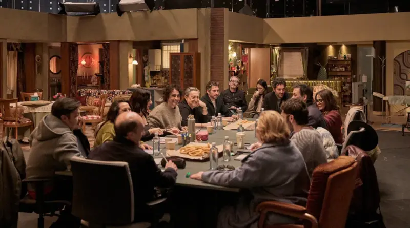 Fotograma de la película 'Aída y vuelta'. Plano general de una reunión del elenco alrededor de una gran mesa llena de papeles, botellas de agua y una caja de porras o churros. Están sentados en lo que parece ser el decorado del salón de la casa de Aída. Los actores (Paco León, Eduardo Casanova, Melani Olivares, etc.) charlan y ríen en un ambiente relajado de trabajo o ensayo.
