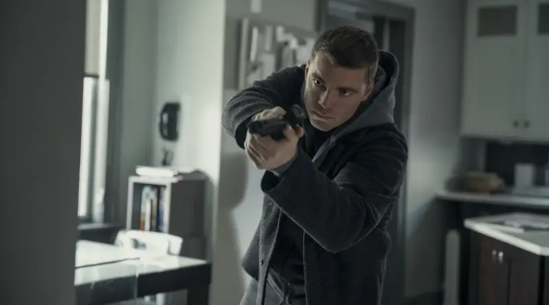 Imagen de la temporada 3 de 'El agente nocturno'. Gabriel Basso, vestido con un abrigo gris de lana, se mueve tácticamente apuntando con su arma hacia una esquina dentro de una habitación moderna y gris.