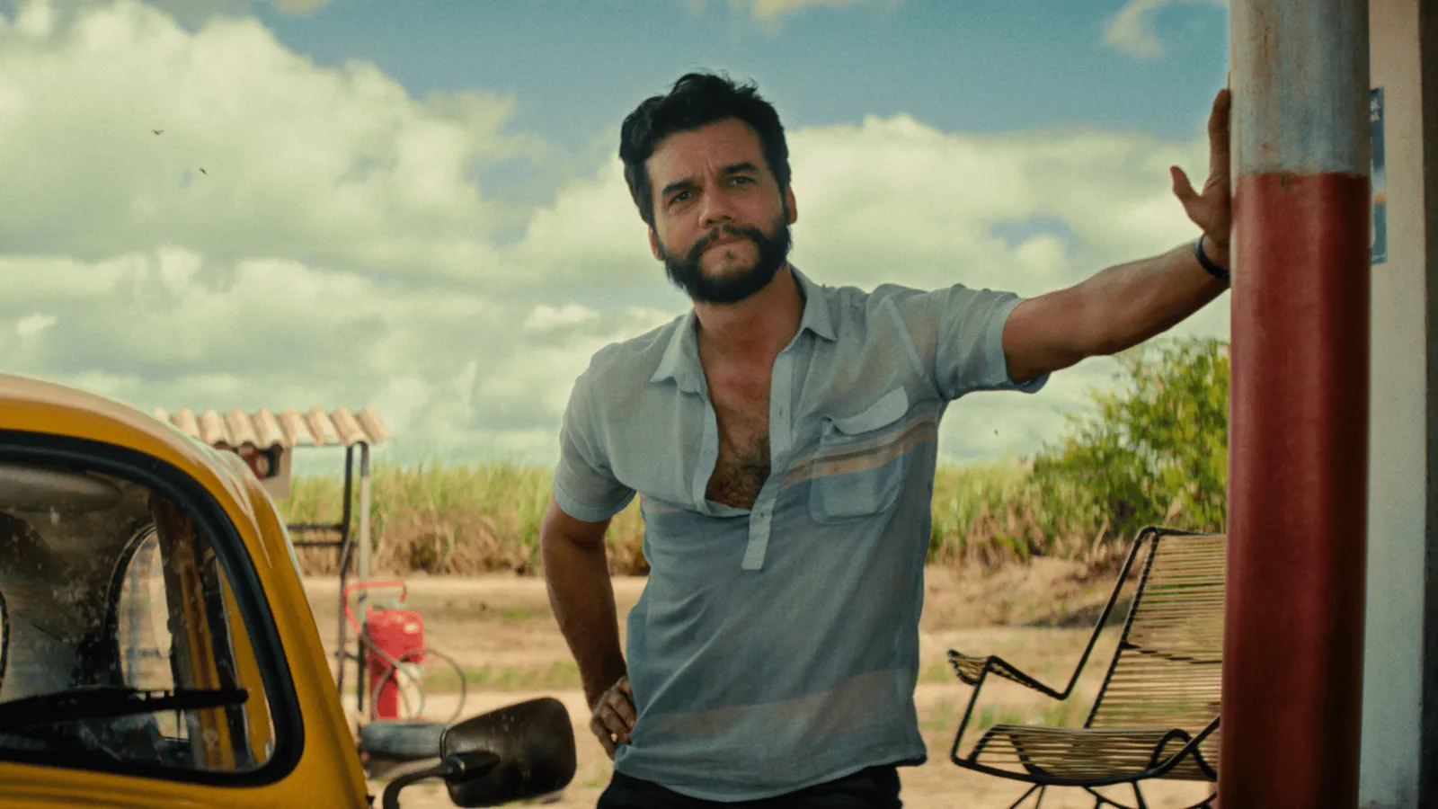 Fotograma de la película 'El Agente Secreto'. El actor Wagner Moura, con barba poblada y cabello oscuro alborotado, se apoya relajadamente con una mano en un poste rojo bajo un cielo azul con nubes. Viste una camisa azul claro de manga corta, desabotonada en el pecho y visiblemente sudada. A su izquierda se ve el morro de un Volkswagen Escarabajo amarillo y, al fondo, un paisaje rural verde con cañaverales.