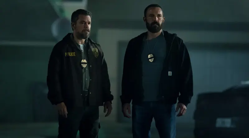 Matt Damon y Ben Affleck en 'El Botín', caracterizados como policías con barba, vistiendo chaquetas oscuras con placas de identificación colgando de sus cuellos, parados uno al lado del otro en una calle oscura de Miami.