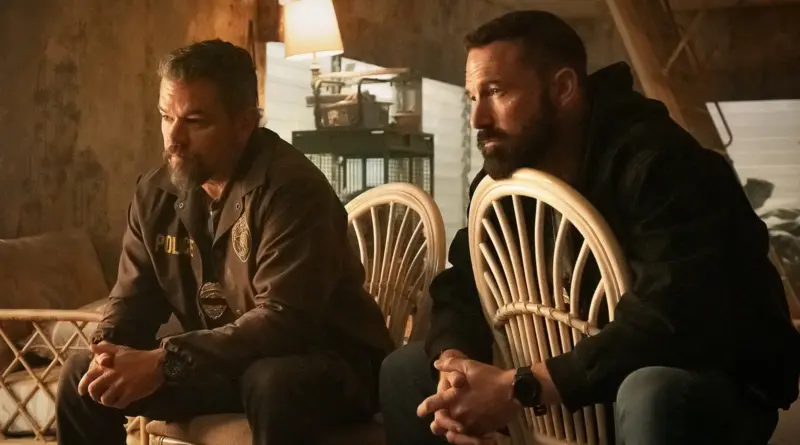 Matt Damon y Ben Affleck sentados en sillas de mimbre en una habitación rústica con luz cálida; ambos lucen barba, expresiones pensativas y llevan placas de policía visibles.