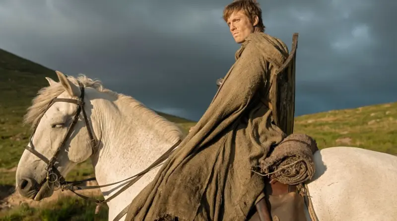 Fotograma de la serie 'El Caballero de los Siete Reinos'. Ser Duncan el Alto (Peter Claffey) montado en un caballo blanco, vistiendo una capa marrón de tela basta y un escudo de madera a la espalda, mirando hacia atrás con expresión seria bajo un cielo nublado y tormentoso en un paisaje verde.