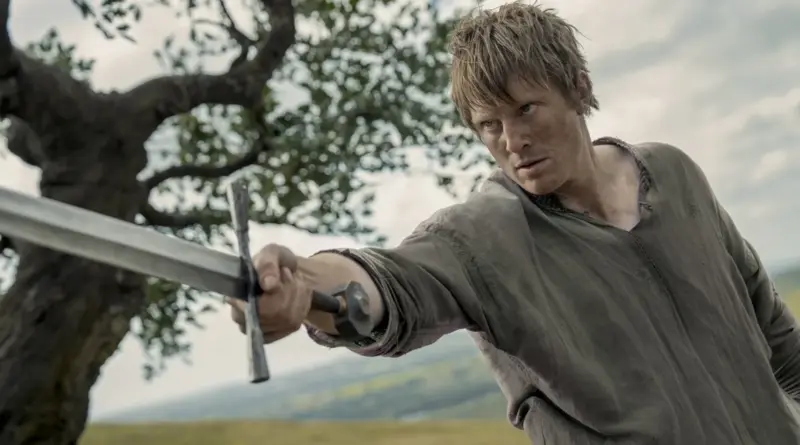 Fotograma de la serie 'El Caballero de los Siete Reinos'. Primer plano de Peter Claffey como Dunk con túnica gris sencilla y la cara manchada de tierra, apuntando firmemente con una espada hacia adelante mientras entrena al aire libre.