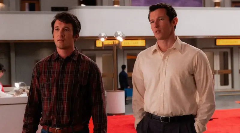 Fotograma de la película 'Eternity'. Miles Teller (izquierda) y Callum Turner (derecha) están de pie en un interior amplio que parece la terminal de un aeropuerto o una oficina gubernamental con moqueta roja. Teller viste una camisa de cuadros roja y negra sobre unos vaqueros, con una expresión de incertidumbre. Turner, a su lado, viste una camisa de color crema y pantalones de vestir oscuros, manteniendo una postura más erguida y formal con las manos en los bolsillos. El fondo muestra una arquitectura moderna de mediados de siglo con luces cálidas.