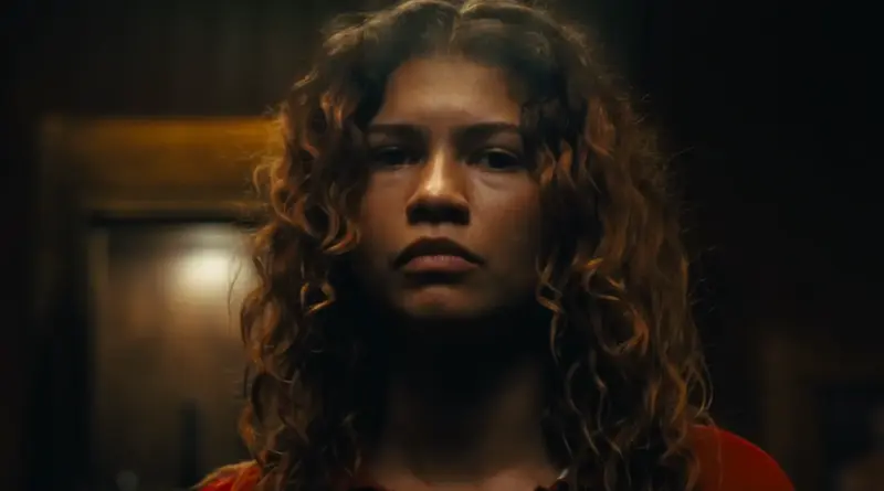 Primer plano frontal de Zendaya con el pelo rizado suelto y una iluminación cálida que resalta su rostro serio y maduro, mirando fijamente al espectador en 'Euphoria.