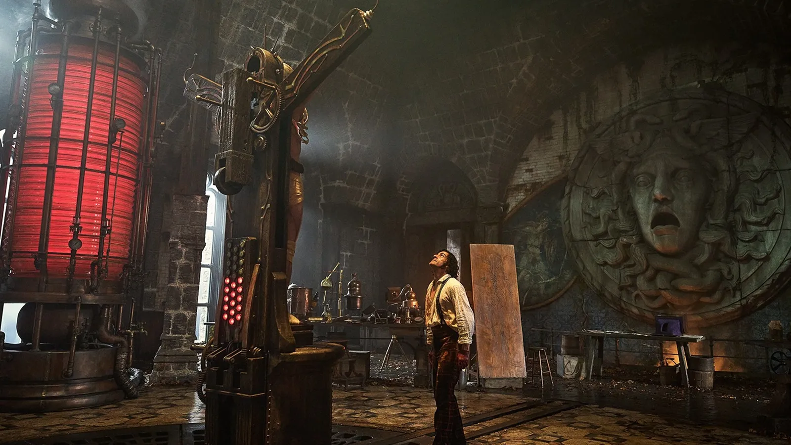 Plano general de un laboratorio gótico industrial con techos abovedados de ladrillo. En el centro, Oscar Isaac (Victor Frankenstein), con camisa blanca y tirantes manchados, mira hacia arriba a un cuerpo vendado sujeto a una compleja máquina vertical de metal y madera con luces rojas. A la izquierda hay un tanque cilíndrico gigante que brilla con luz roja intensa. En la pared de la derecha, domina un enorme relieve de piedra de la cabeza de Medusa con la boca abierta en un grito.