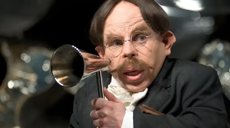 Primer plano de Warwick Davis en las películas de 'Harry Potter' caracterizado como el Profesor Filius Flitwick, con bigote rubio, gafas redondas y sosteniendo una trompetilla plateada para la audición frente a un fondo desenfocado.