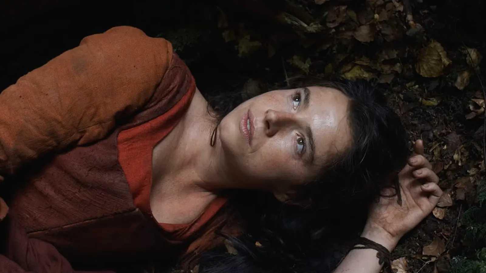 Primer plano en ángulo picado de la actriz Jessie Buckley (interpretando a Anne Hathaway) en 'Hamnet', tumbada sobre un suelo de tierra oscura cubierto de hojas secas y vegetación. Viste un vestido de época de tela rústica en color teja o rojizo. Tiene la mirada fija hacia el cielo con una expresión de vulnerabilidad, asombro o dolor contenido, y los ojos vidriosos.