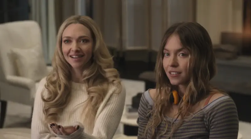 Crítica de 'La asistenta': Sydney Sweeney se echa a la espalda el best seller del año 1 Primer plano de Amanda Seyfried (izquierda) y Sydney Sweeney (derecha) sentadas en una habitación iluminada suavemente en una escena de la película 'La asistenta'. Amanda, con el cabello rubio y ondulado, viste un jersey blanco de punto y sonríe con amabilidad mientras extiende su mano derecha en un gesto explicativo. Sydney, con el cabello castaño claro y liso, viste una camiseta de rayas horizontales y lleva unos auriculares naranjas y negros alrededor del cuello, mostrando una expresión de curiosidad o asombro. El fondo está ligeramente desenfocado, dejando ver mobiliario moderno.