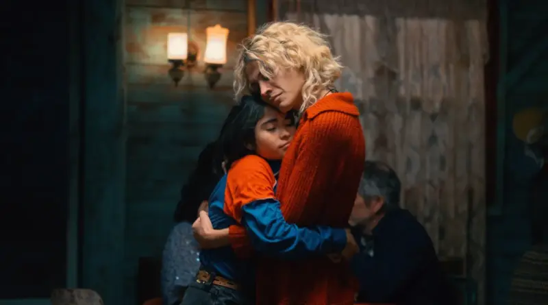 Fotograma de la película 'La misteriosa mirada del flamenco'. Una mujer con cabello rubio platino ondulado y un jersey de punto naranja abraza con fuerza y dolor a una niña de pelo oscuro que viste una camiseta azul y naranja. La niña tiene los ojos cerrados, descansando en el pecho de la mujer. El fondo es un interior doméstico con luz tenue y cortinas de encaje.