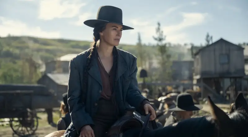 Fotograma de la serie 'Los abandonados'. Plano medio de Lena Headey montada a caballo bajo la luz del día, vistiendo un sombrero negro de ala ancha y mirando hacia el horizonte con expresión estoica, con edificios de madera de un pueblo al fondo.