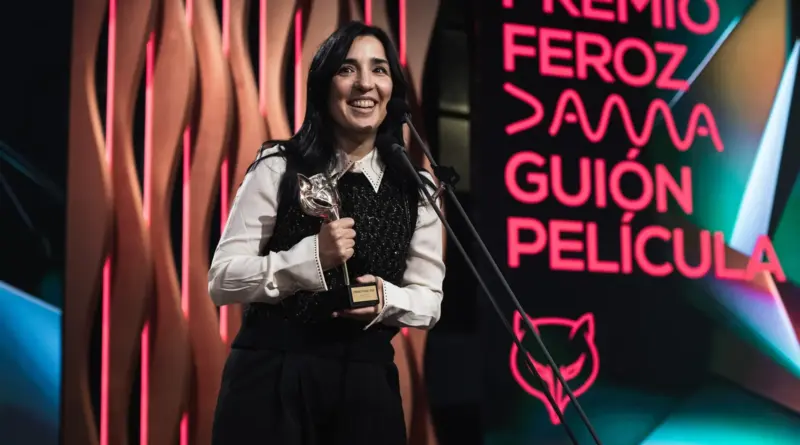 Plano medio de Alauda Ruiz de Azúa, una mujer de cabello oscuro sonriendo ampliamente frente a un micrófono, en los Premios Feroz 2026. Sostiene con ambas manos el trofeo Feroz (una máscara de zorro dorada) contra su pecho. Viste una camisa blanca de cuello picudo bajo un chaleco negro texturizado. A su derecha, una pantalla digital muestra en letras de neón rosa y rojo: "PREMIO FEROZ DAMA GUIÓN PELÍCULA".