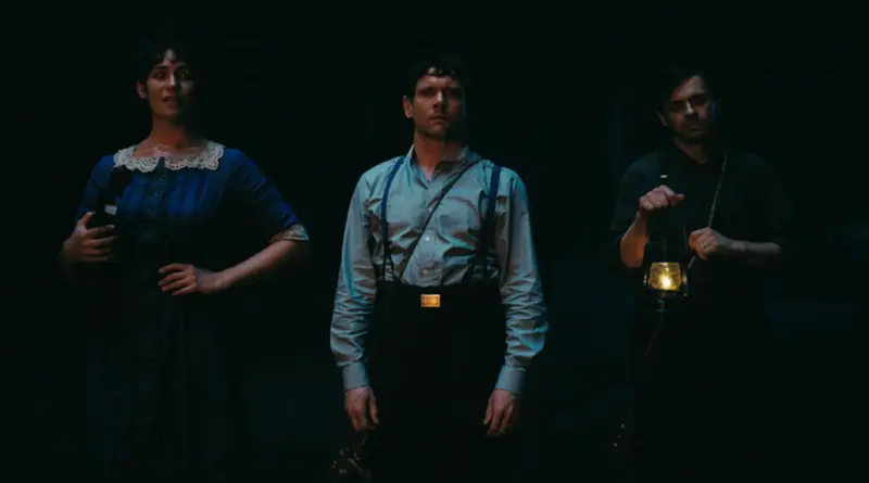 Fotograma de la película 'Sinners' (Los Pecadores) con tres personajes de pie en la oscuridad. En el centro, Jack O'Connell viste camisa gris y tirantes, mirando con expresión de shock o intensidad.