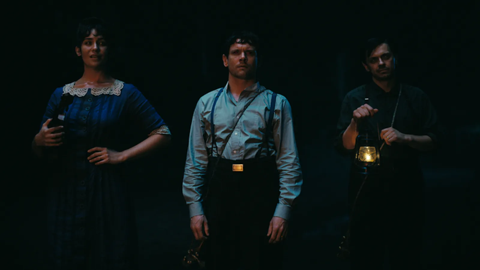 Fotograma de la película 'Sinners' (Los Pecadores) con tres personajes de pie en la oscuridad. En el centro, Jack O'Connell viste camisa gris y tirantes, mirando con expresión de shock o intensidad.